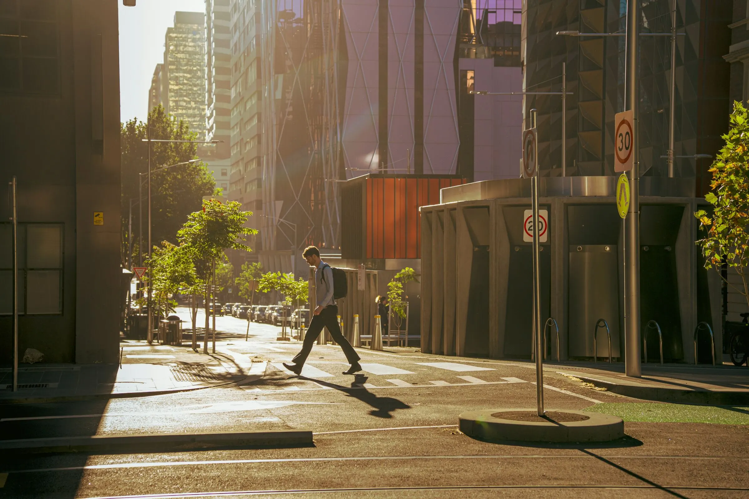 A man with a backpack crosses a sunlit city street